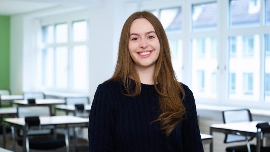 A young woman with long brown hair and a dark sweater smiles as she stands in a bright classroom with large windows, empty desks, and MaxBrain learning materials in the background.