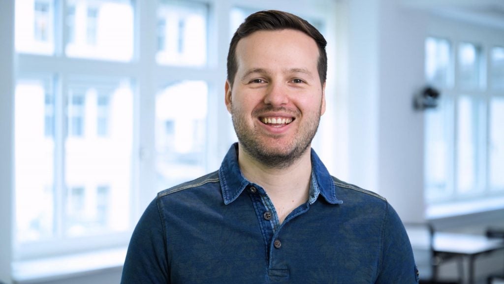 A man with short brown hair and a beard, wearing a blue button-up shirt, smiles as he stands in a bright room with large windows—the perfect setting for productive MaxBrain sessions.