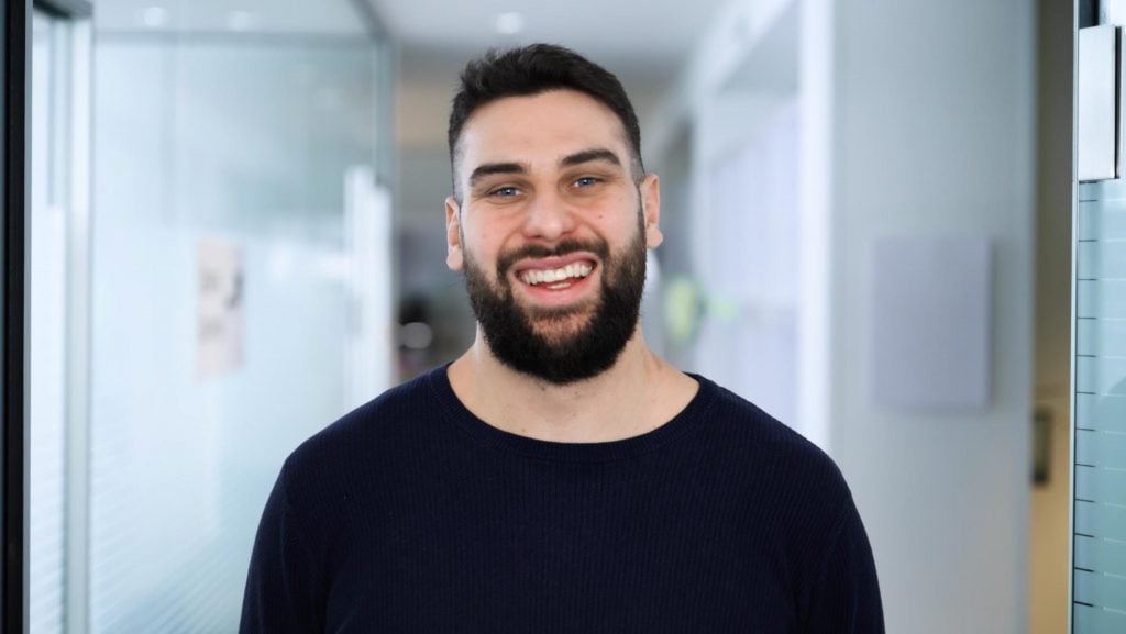 A bearded man with dark hair and a dark sweater is standing in a bright, modern hallway at MaxBrain, smiling at the camera. The background consists of blurred glass walls and soft lighting.