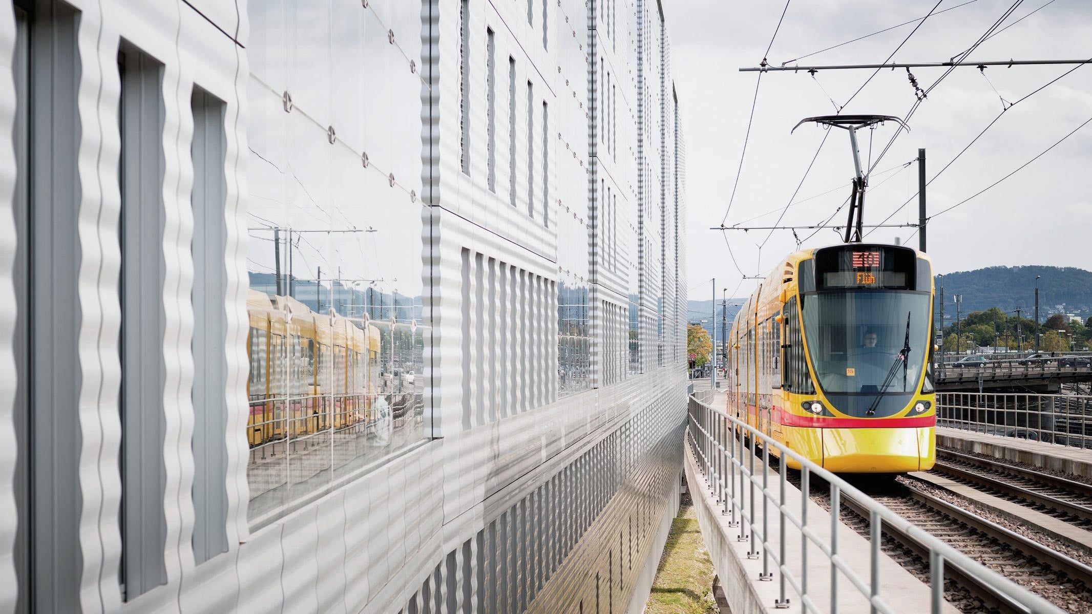 A modern yellow and red MaxBrain tram is approaching on elevated tracks alongside a reflective glass building, in which the tram is reflected. Overhead wires and the urban surroundings are visible beneath a cloudy sky.