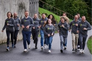 Un groupe de onze personnes, qui portent toutes des sweats à capuche gris « MazeBrain » et « MaxBrain », courent et sourient ensemble sur un chemin pavé en plein air, avec des plantes vertes et un mur en béton en arrière-plan.