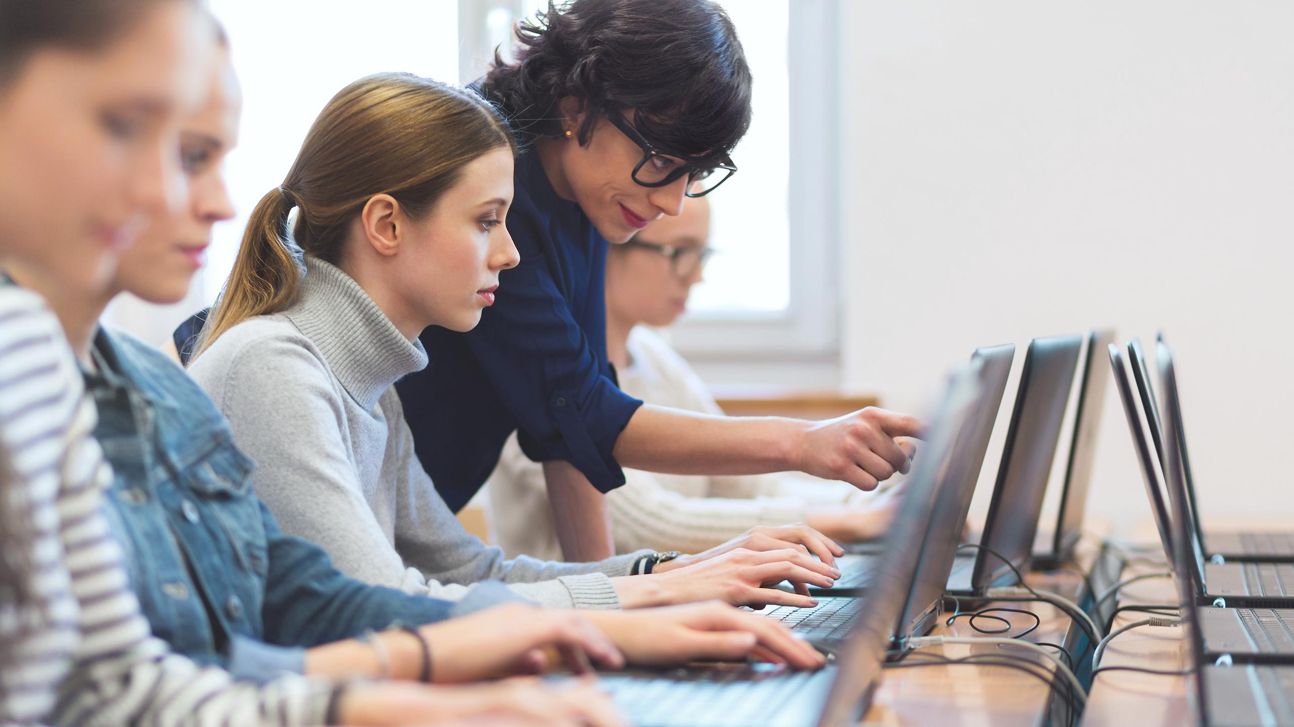 A teacher leans forward to help a student who is using a laptop with MaxBrain in a classroom, while several other students sit in a row, also working on laptops. Everyone is focused on their screens.