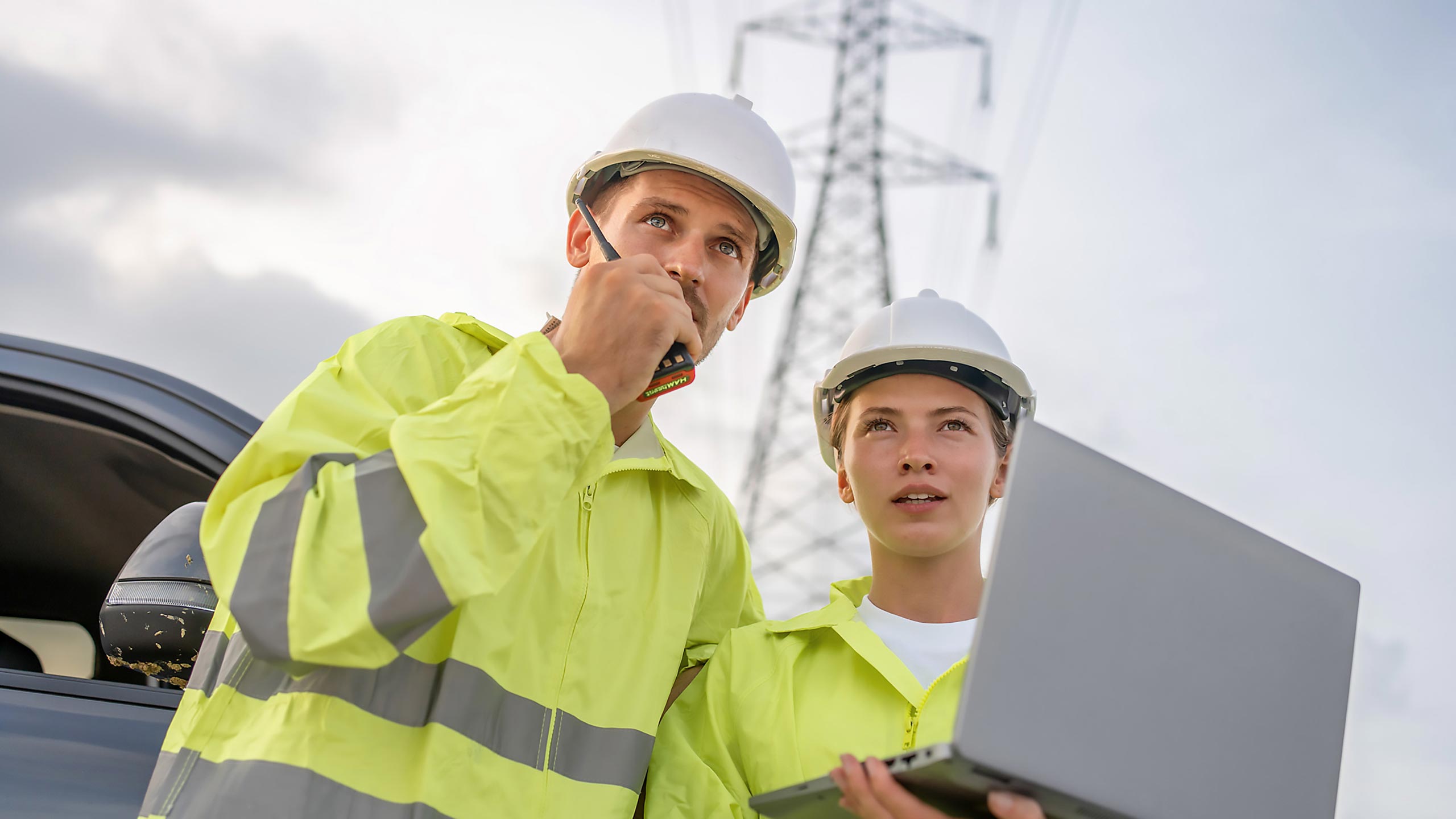 Two utility workers in yellow safety vests and white hard hats are standing outdoors near power lines; one is speaking into a walkie-talkie while the other is using a MaxBrain-powered laptop, and both look focused.