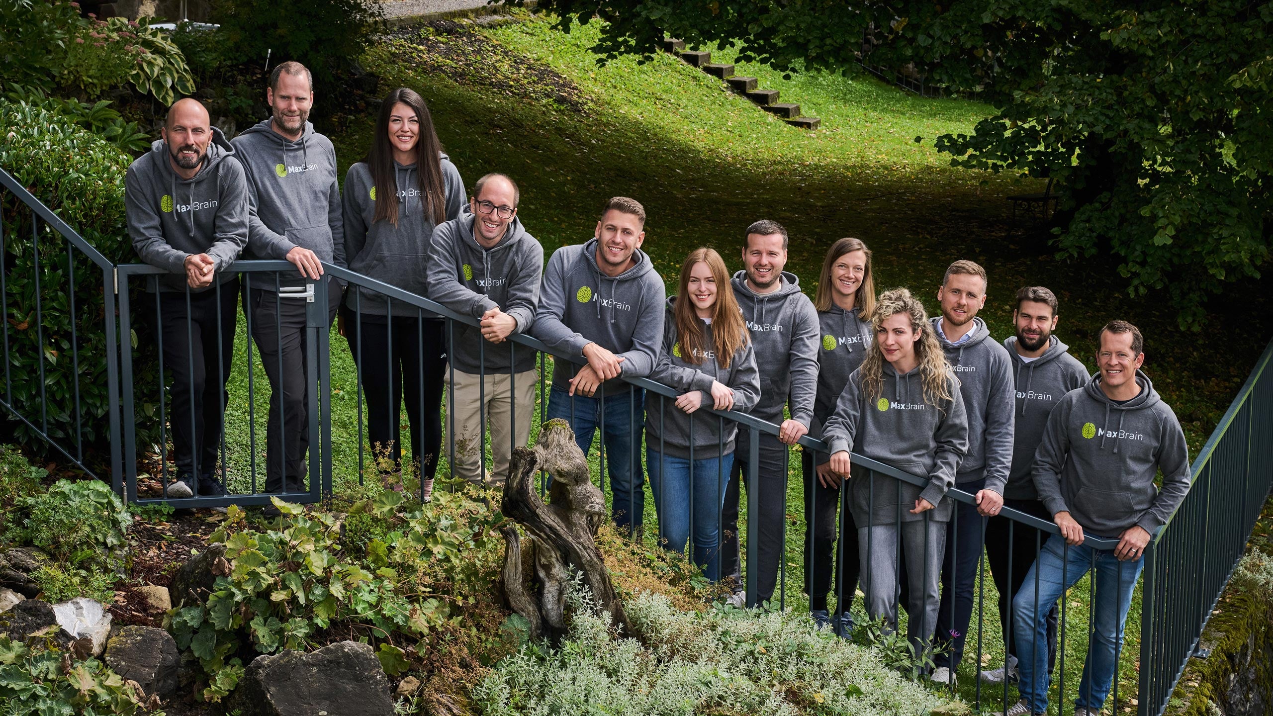 Twelve people wearing matching gray MaxBrain hoodies with a green logo are standing and leaning against a black railing outdoors, surrounded by greenery and plants, and smiling at the camera.